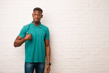 Genç Afro-Amerikan siyahi adam şaşkın, şaşkın ve güvensiz hissediyor. Kendini merak edip bana mı soruyor? tuğla duvara karşı