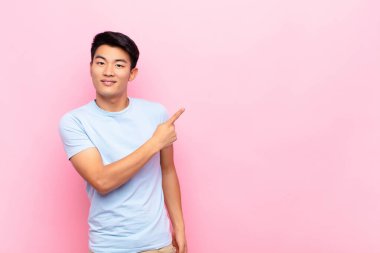 Genç Çinli adam heyecanlı ve şaşırmış görünüyor. Yana ve yukarıya doğru düz renkli duvara uzayı gösteriyor.