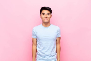 Genç Çinli adam mutlu ve şapşal görünüyor. Geniş, eğlenceli, kaçık bir gülüşü ve düz renkli duvarları olan gözleri var.