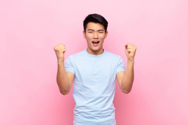 Genç Çinli adam mutlu, şaşırmış ve gururlu hissediyor, bağırıyor ve başarıyı kutluyor düz renkli duvara karşı büyük bir gülümsemeyle.