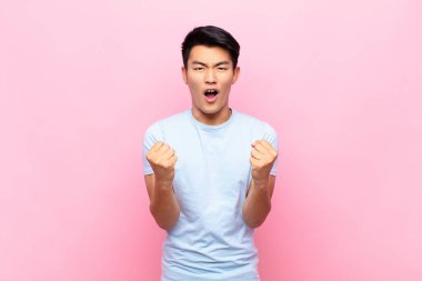 Genç Çinli adam sinirli, sinirli, sinirli bakışlar ve sıkı yumruklarla bağırıyor, düz renkli duvara karşı öfkeli hissediyor.