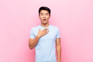 Genç Çinli adam şok olmuş, şaşırmış ve şaşırmış hissediyor, elleri göğsünde ve ağzı açık, kim diyor, ben mi? yassı renk duvarına karşı