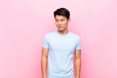 Genç Çinli bir adam üzgün, üzgün ya da kızgın ve olumsuz bir tavır takınıyor, düz renk duvarına karşı anlaşmazlık içinde.