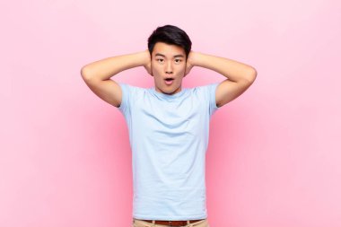 Genç Çinli adam heyecanlı ve şaşırmış görünüyor, ağzı açık, iki eli başının üstünde, düz duvara karşı şanslı bir kazanan gibi hissediyor.