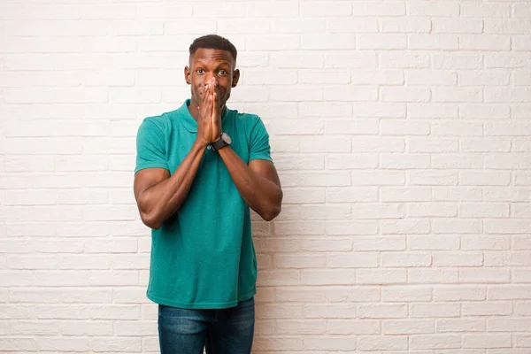 young african american black man happy and excited, surprised and amazed covering mouth with hands, giggling with a cute expression against brick wall