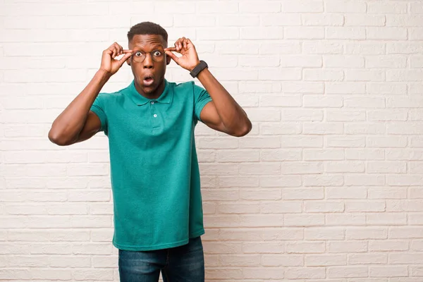 young african american black man feeling shocked, amazed and surprised, holding glasses with astonished, disbelieving look against brick wall