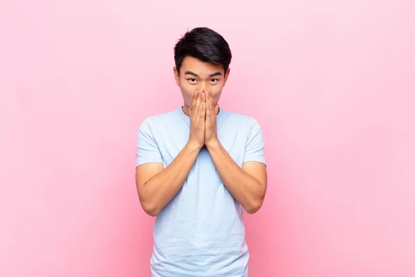 young chinese man happy and excited, surprised and amazed covering mouth with hands, giggling with a cute expression against flat color wall