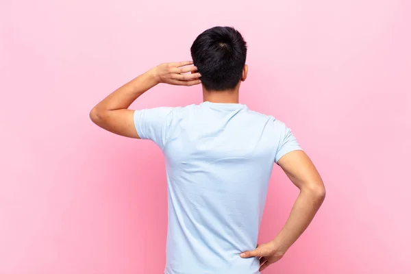 young chinese man thinking or doubting, scratching head, feeling puzzled and confused, back or rear view against flat color wall