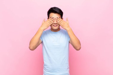 Genç Çinli adam gülümsüyor ve mutlu hissediyor, iki eliyle gözlerini kapatıyor ve düz renkli duvara karşı inanılmaz bir sürpriz bekliyor.