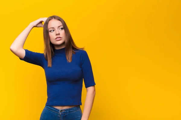 young red head woman feeling puzzled and confused, scratching head and looking to the side against flat wall