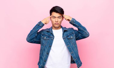 Ciddi ve konsantre görünümlü genç Çinli bir adam düz renk duvarına karşı beyin fırtınası yapıyor ve kafa yoruyor.