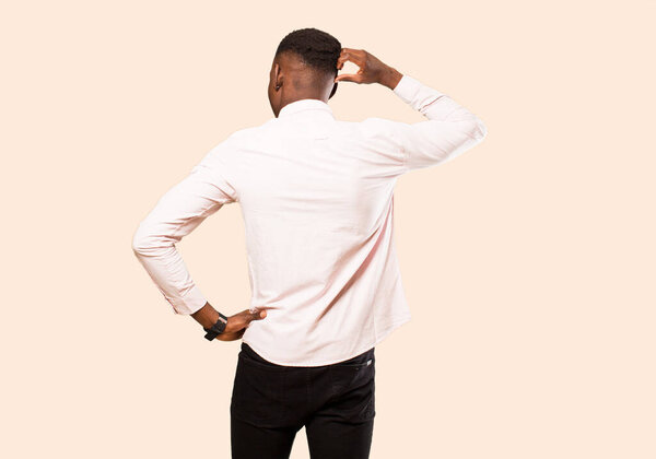 Young african american black man feeling clueless and confused, thinking a solution, with hand on hip and other on head, rear view against beige wall