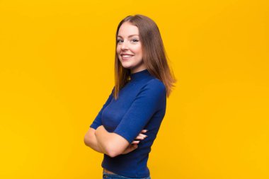 Genç kızıl saçlı kadın kameraya çapraz kollarıyla gülümsüyor ve mutlu, kendinden emin, tatmin olmuş bir yüz ifadesi düz duvara karşı yanal bir bakış.