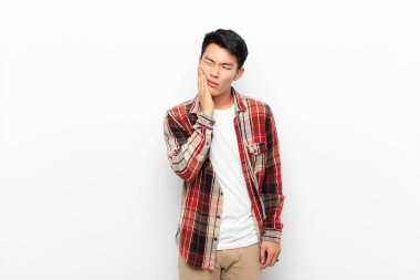 Genç Çinli adam yanağını tutuyor ve acı çekiyor, hasta, sefil ve mutsuz hissediyor, düz renkli duvara karşı bir dişçi arıyor.