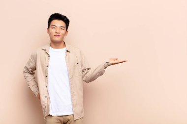 Genç Çinli adam gülümsüyor, kendinden emin, başarılı ve mutlu hissediyor, fotokopi odasında düz renkli duvara karşı konsept veya fikir gösteriyor.