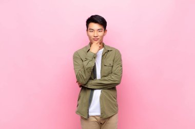 Genç Çinli adam mutlu ve gülümsüyor, çenesi üzerinde, merak ediyor ya da soru soruyor, düz renkli duvarla seçenekleri karşılaştırıyor.