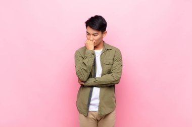 Genç Çinli adam ciddi, düşünceli ve endişeli, elleri çenesine yaslanmış bir şekilde düz duvara bakıyor.