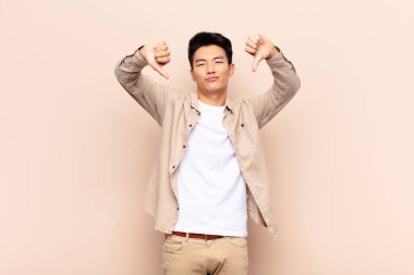 Genç Çinli adam üzgün, hayal kırıklığına uğramış ya da kızgın görünüyor. Anlaşmazlıkta baş parmağını gösteriyor. Düz renkli duvara karşı hayal kırıklığına uğramış hissediyor.