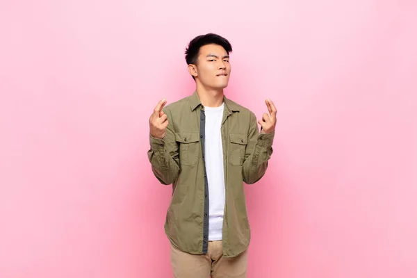 young chinese man crossing fingers anxiously and hoping for good luck with a worried look against flat color wall