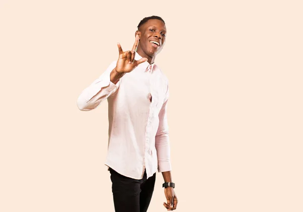 young african american black man feeling happy, fun, confident, positive and rebellious, making rock or heavy metal sign with hand against beige wall