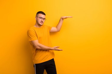 Genç yakışıklı bir adam iki eli de yan tarafında fotokopi alanı olan bir nesneyi tutuyor, düz arkaplana karşı bir nesneyi gösteriyor, sunuyor ya da reklamını yapıyor.