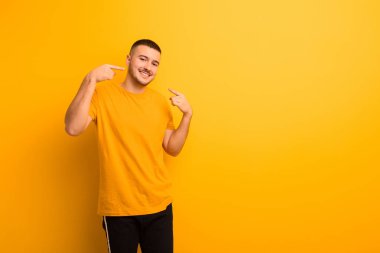 Genç ve yakışıklı bir adam kendi gülüşüne güvenerek gülümsüyor, pozitif, rahat, düz arkaplan karşısında tatmin olmuş bir tavır sergiliyor.