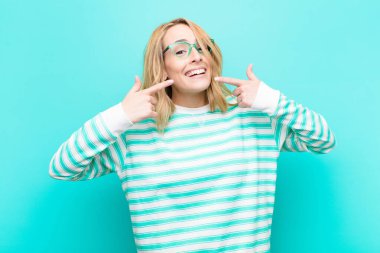 Genç, güzel sarışın kadın, kendine güvenerek gülümsüyor. Geniş gülüşünü gösteriyor. Olumlu, rahat, tatmin olmuş, düz renkli duvara karşı tavır alıyor.