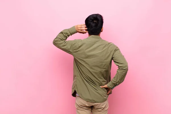 young chinese man thinking or doubting, scratching head, feeling puzzled and confused, back or rear view against flat color wall