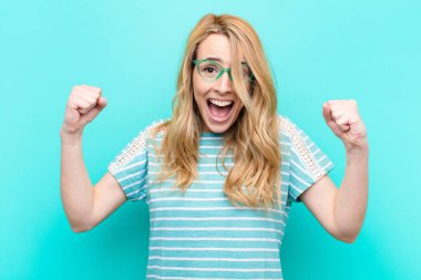Genç, güzel, sarışın bir kadın mutlu, şaşırmış ve gururlu, bağırıyor ve başarıyı kutluyor. Düz renkli duvara karşı kocaman bir gülümsemeyle.