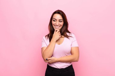 Genç, güzel bir kadın mutlu, kendine güvenen bir ifadeyle gülümsüyor. Elleri çenesinde, pembe duvara bakıyor ve merak ediyor.