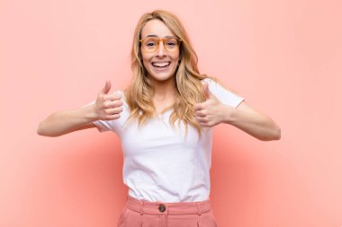 young pretty blonde woman smiling broadly looking happy, positive, confident and successful, with both thumbs up against flat color wall clipart