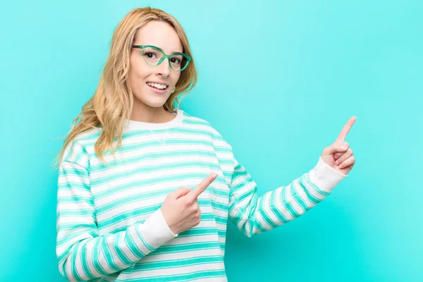 young pretty blonde woman smiling happily and pointing to side and upwards with both hands showing object in copy space against flat color wall