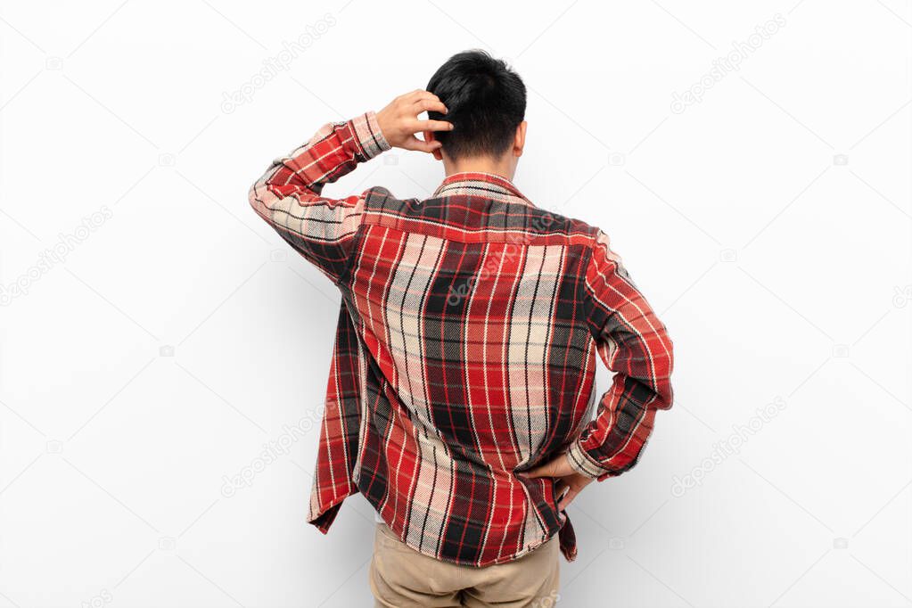 young chinese man feeling clueless and confused, thinking a solution, with hand on hip and other on head, rear view against flat color wall