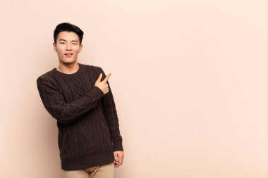 Genç Çinli adam heyecanlı ve şaşırmış görünüyor. Yana ve yukarıya doğru düz renkli duvara uzayı gösteriyor.