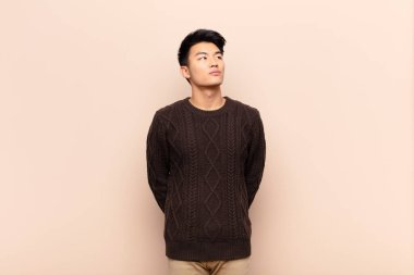 Endişeli, kafası karışık, cahil bir ifadeyle genç Çinli adam, kopya alanı arıyor, düz renkli duvara karşı şüpheyle bakıyor.