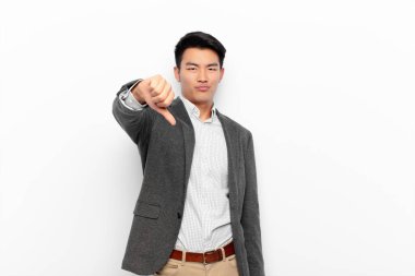young chinese man feeling cross, angry, annoyed, disappointed or displeased, showing thumbs down with a serious look against flat color wall clipart