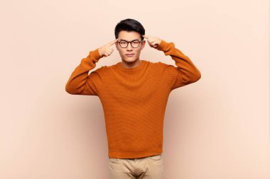 Ciddi ve konsantre görünümlü genç Çinli bir adam düz renk duvarına karşı beyin fırtınası yapıyor ve kafa yoruyor.