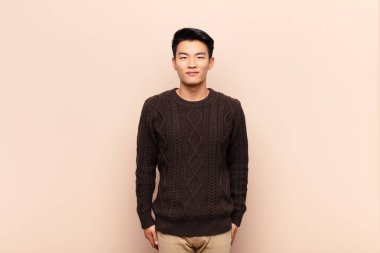 Genç Çinli adam pozitif ve kendinden emin gülümsüyor. Tatmin olmuş, arkadaş canlısı ve mutlu görünüyor. Düz renkli duvara karşı.