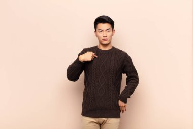 Genç Çinli adam mutlu, gururlu ve şaşırmış görünüyor, neşeli bir şekilde kendini gösteriyor, kendinden emin ve düz renkli duvara karşı yüce hissediyor.