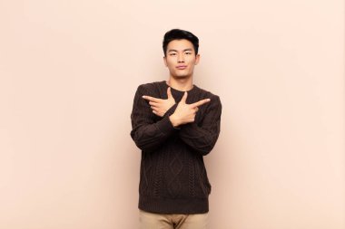 Genç Çinli adam şaşkın ve şaşkın görünüyor, güvensiz ve zıt yönleri işaret ediyor düz renkli duvara karşı şüpheyle