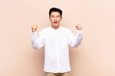 Genç Çinli adam mutlu, şaşırmış ve gururlu hissediyor, bağırıyor ve başarıyı kutluyor düz renkli duvara karşı büyük bir gülümsemeyle.