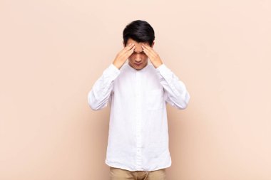 Genç Çinli bir adam stresli ve hayal kırıklığına uğramış görünüyor. Baskı altında, baş ağrısı ile çalışıyor ve düz renkli duvarlara karşı sorunları var.