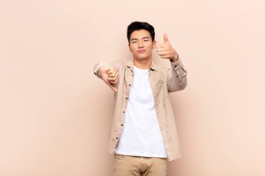 Genç Çinli bir adam kafası karışmış, bilgisiz ve kararsız hissediyor, farklı seçeneklerde iyi ve kötü şeyleri tartıyor ya da düz renkli duvarlara karşı seçenekleri tartıyor.