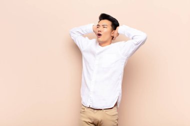 Ağzı açık genç Çinli bir adam, korkunç bir hata yüzünden dehşete düşmüş ve şok olmuş görünüyor, düz renkli duvara karşı ellerini kaldırıyor.