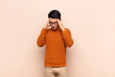 Genç Çinli bir adam stresli ve hayal kırıklığına uğramış görünüyor. Baskı altında, baş ağrısı ile çalışıyor ve düz renkli duvarlara karşı sorunları var.