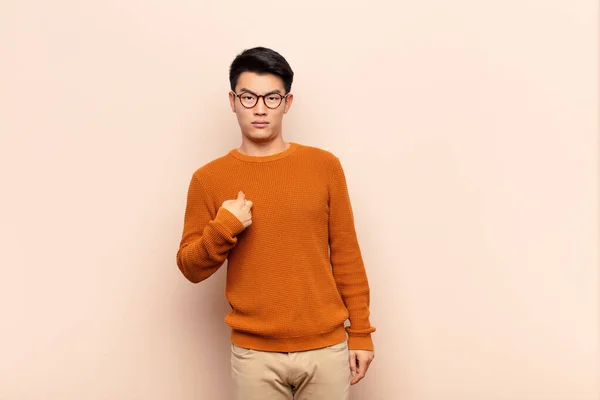 young chinese man feeling confused, puzzled and insecure, pointing to self wondering and asking who, me? against flat color wall