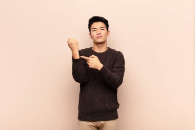 Genç Çinli adam sabırsız ve kızgın görünüyor, saati gösteriyor, dakiklik istiyor, düz renkli duvara karşı zamanında olmak istiyor.