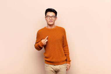 Genç Çinli adam gururlu, kendinden emin ve mutlu görünüyor, gülümsüyor ve kendini işaret ediyor ya da düz renkli duvara karşı bir numara işareti yapıyor.