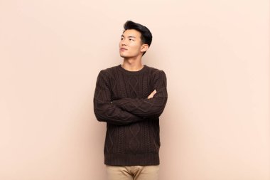 Genç Çinli adam mutlu, gururlu ve umutlu hissediyor, merak ediyor ya da düşünüyor, düz renkli duvara karşı çapraz kollarıyla boşluğu dolduruyor.