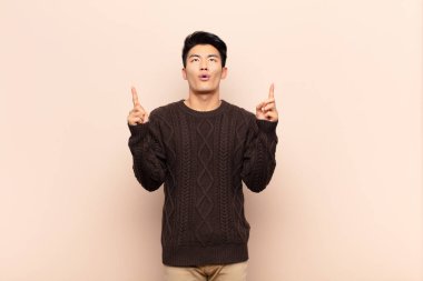 Genç Çinli adam şaşırmış, şaşırmış ve ağzı açık görünüyor, iki eliyle yukarıya bakıyor düz renkli duvara uzayı kopyalamak için.